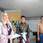 A woman in a pink jacket gives a speech into a microphone, watched in the background by a man and a woman in traditional Thai dress.