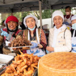 Four Thai women at the food stall, one holds a large tray of skewered meat. In the ofreground is a tray piled with glazed chicken wings and a large covered bamboo basket.
