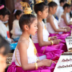 A young girl playing a traditional Thai stringed instrument with a pair of hammers as part of a larger orchestra.